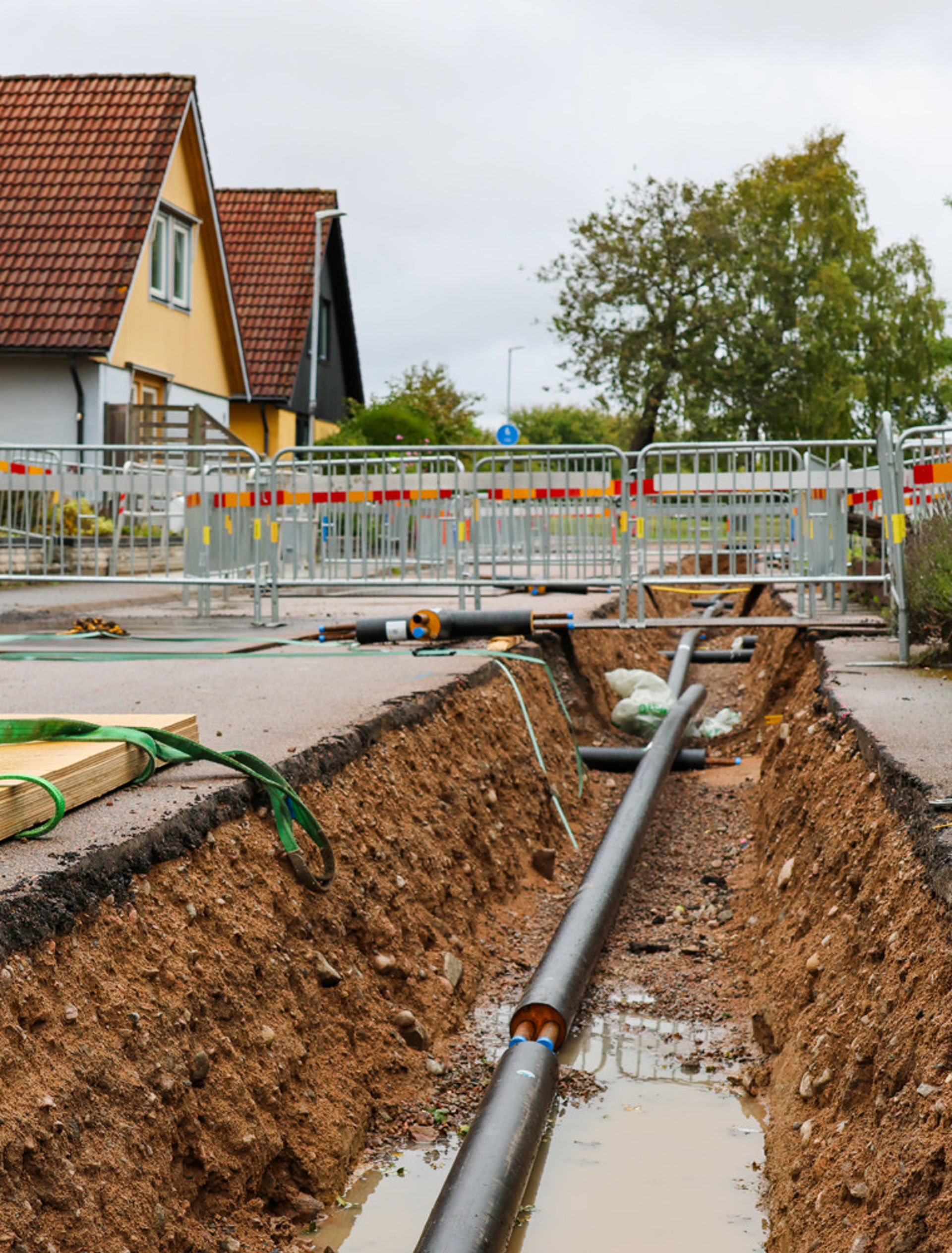 En schakt i en gata med pågående bygg-/anläggningsarbete – synliga rör ligger ner i schaktet, jord och grus runt omkring, säkerhetsstaket och trafikavspärrning i bakgrunden, samt bostadshus och träd bakom arbetsplatsen.